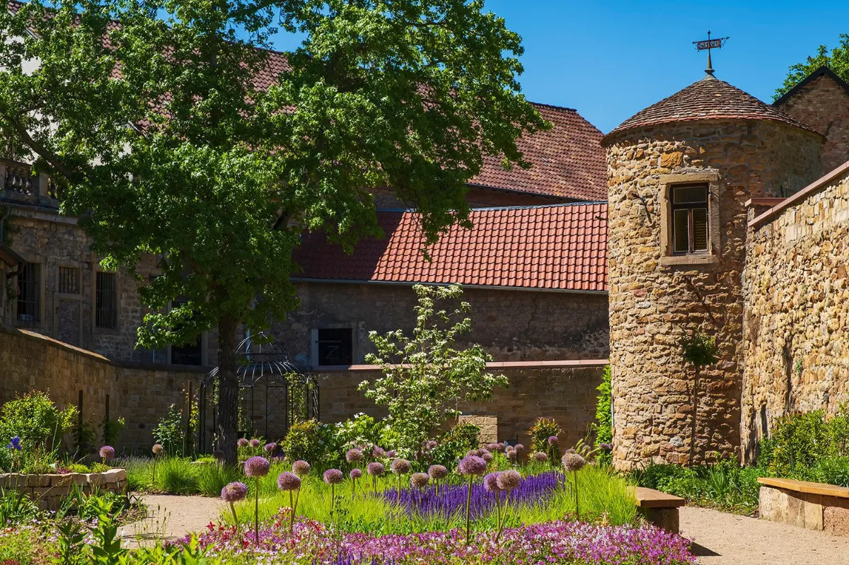 Historischer Turmschreiberturm in Deidesheim mit liebevoll angelegtem Garten und alter Stadtmauer bei Sonnenschein.